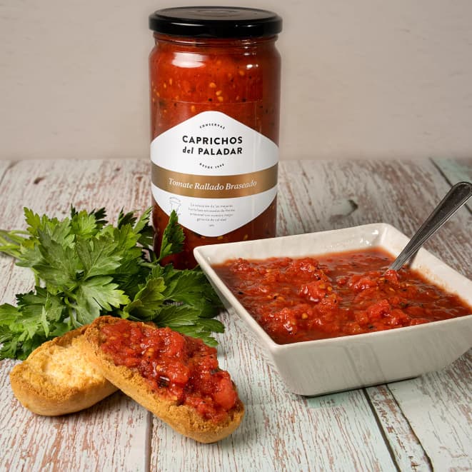 Jar of Caprichos del Paladar tomato relish with a bowl of relish, bread, and parsley on a wooden surface.